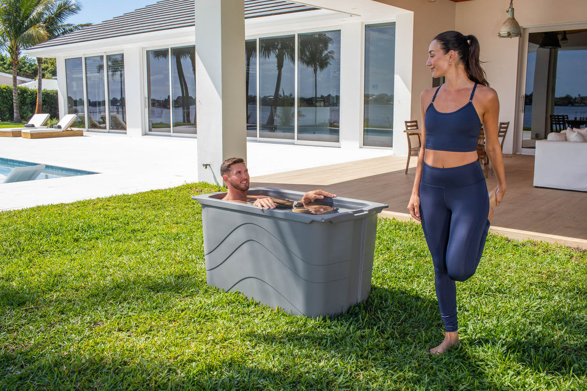 Man using a grey Frozin cold plunge tub beside a woman in a landscaped backyard